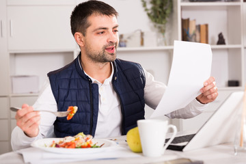 Young man eating vegetable salad and working at laptop