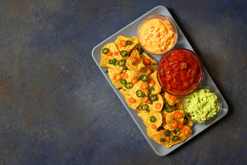 Mexican yellow corn tortilla chips nachos with jalapeno, guacamole, cheese sauce and salsa on dark blue background