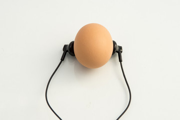 small black headphones lie next to the chicken egg as if the egg is listening to music, white...