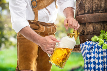 Bavarian man in leather trousers is pouring a large lager beer in tap from wooden beer barrel