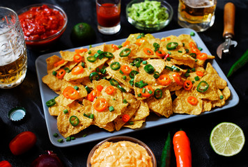 Mexican yellow corn tortilla chips nachos with jalapeno, guacamole, cheese sauce and salsa on black background. Glasses with beer, lime, chili peppers, cherry tomatoes