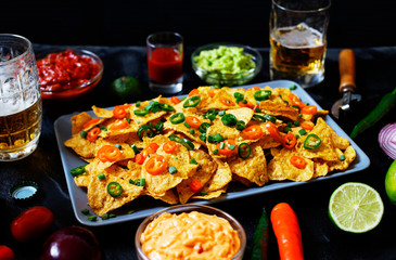 Mexican yellow corn tortilla chips nachos with jalapeno, guacamole, cheese sauce and salsa on black background. Glasses with beer, lime, chili peppers, cherry tomatoes