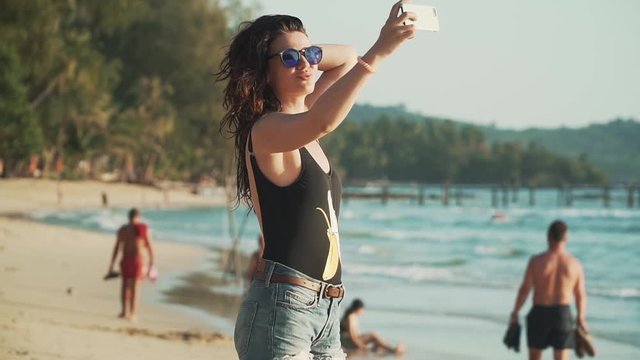 Pretty young woman in sunglasses and swimsuit takes selfie on beautiful sandy beach by sea. Happy smiling girl pulls her hair, spinning and taking pictures of herself with smartphone. Slow motion.