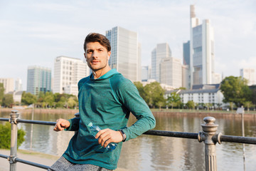 Photo of serious man holding plastic bottle and using earphones while bending on railing near city...