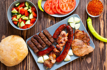 Traditional Serbian and Balkan grilled meat called mesano meso. Balkan barbeque (rostilj) served with Serbian salad, hot peppers, bread, tomato, onions, and paprika powder. Wooden background. Top view