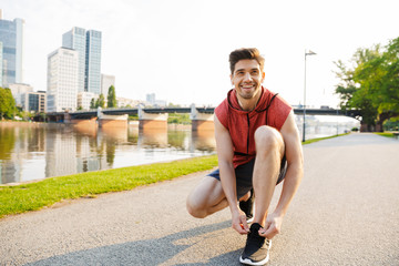 Photo of happy man using earphones and tying his shoelaces while working out near city riverfront