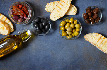 Different types of olives, bruschetta, sun-dried tomatoes and olive oil. Mediterranean snacks. Top view. Dark background. Space for text