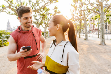 Photo of sporty pleased man and woman using cellphone and talking while working out in city boulevard