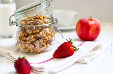 Homemade granola in a glass jar on a light background. Ingredients for a healthy breakfast - granola, apple, strawberry and milk. Close-up