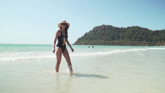 Young caucasian woman walks along sandy beach by the sea. Girl in swimsuit and straw hat enjoys vacation on paradise tropical island with sandy beach, calm turquoise sea and green trees on the shore.