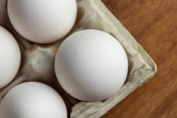 Chicken eggs are raw in the tray. The shell is white in eggs. In one section is an egg shell. On one egg lies a feather of white color. Wooden background. View from above. Close-up