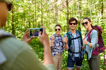 travel, tourism and hike concept - group of friends with backpacks being photographed by smartphone in forest