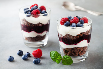Layered trifle dessert with chocolate sponge cake, whipped cream, berries and fruit jelly in serving glasses. - obrazy, fototapety, plakaty