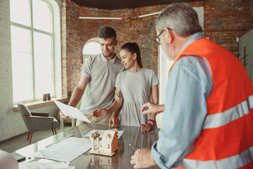 Foreman or achitect engineer shows future house, office or store design plans and model to a young couple. Meeting at the construction office to talk about facade, interior decoration, home layout.