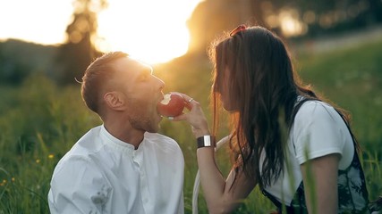 A young man and woman are sitting on a green meadow and eating an apple. Romance, love