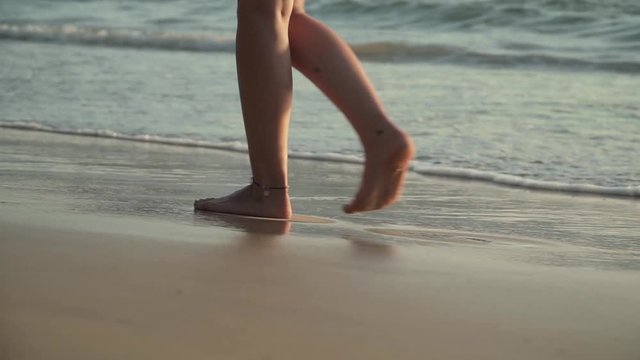Close-up of legs of young woman walking along tropical beach at sunset. Foamy sea waves gentrly wash sand and feet, warm soft evening sun reflects on wet surface. Slow motion footage.