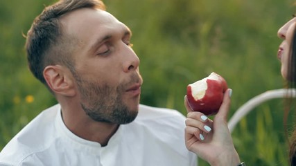 A young man and woman are sitting on a green meadow and eating an apple. Romance, love