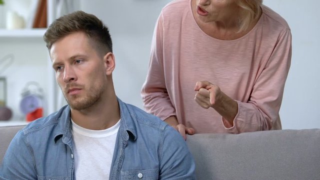 Mature mother nagging at irritated adult son sitting on sofa, family conflict