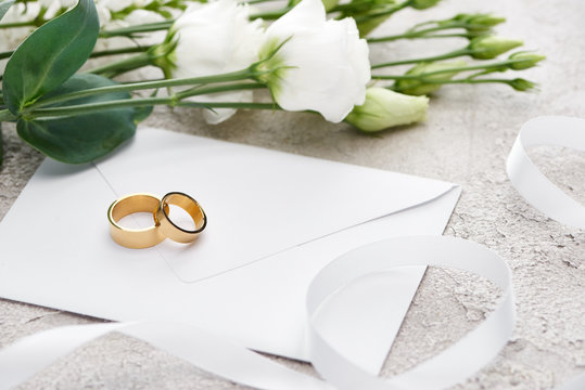 golden wedding rings on white envelope near eustoma flowers and white ribbon