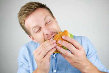 young handsome man biting fresh tasty burger from fast food restaurant looks hungry eating on isolated white background