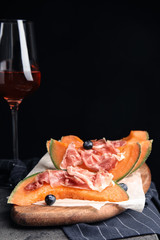 Wooden board with melon, prosciutto and blueberries on table against black background