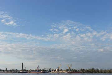 Industrial landscape, cranes, ships and factories on the river bank, against the blue sky.