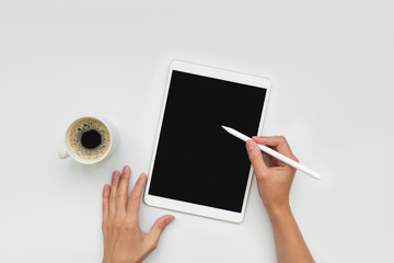 Cup of coffee and feminine hands drawing on tablet by pencil on white desk. Top view. Copy space. Mock-up