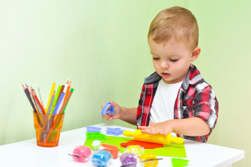 Happy boy playing with plasticine. A two-year-old baby boy in a red checked shirt sits at a table