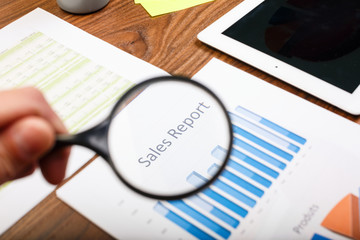 Hand holds magnifying glass over paper with sales report.