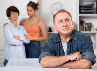 Woman and her daughter are sympathying their sad father who is sitting at the table