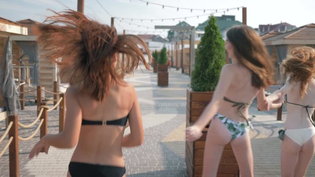 rest with friends, young slender girlfriends in swimsuits have fun on vacation by the sea, running along a luxurious promenade against the backdrop of a bungalow for relaxation, rear view