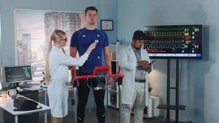 Female cardiology technologist listening to athlete heartbeat with a stethoscope while her colleague learning sportsman cardiogram. In Scientific Sports Laboratory. - Powered by Adobe