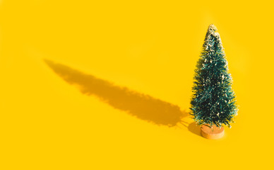Miniature toy artificial Pine tree on wooden base on bright yellow Christmas background with long shadow. Concept of New Year, Christmas holiday greeting card.