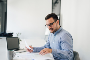 Fototapeta premium Happy young businessman looking at papers graphs and charts using laptop at his office desk