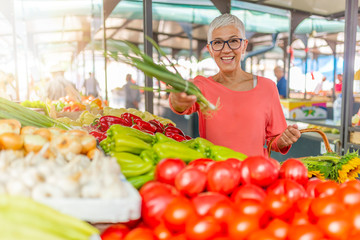 Senior woman buying vegetables at farmers market. Woman picking fresh produce at the market. Senior Woman shopping at an outdoor market. Woman shopping on the farmer's market
