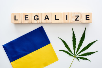 Fototapeta na wymiar top view of green cannabis leaf and legalize lettering on wooden cubes near flag of Ukraine on white background
