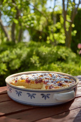 Vertical view to fruit pie in rustic ceramic form on red painted wooden board with green trees in the background. 