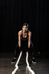 Fototapeta na wymiar selective focus of beautiful sportswoman exercising with battle ropes on black