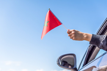 Boy holding Morocco Flag from the open car window on the sky background. Concept