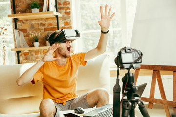 Young caucasian male blogger with professional equipment recording video review of VR glasses at home. Blogging, videoblog, vlogging. Man using virtual reality headset while streaming live.