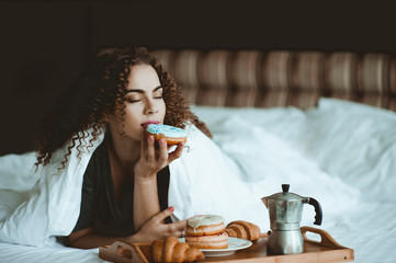 Beautiful blonde woman eating donuts lying in bed in hotel room closeup. Good morning. Breakfast.