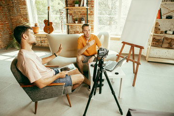 Two young caucasian male bloggers in casual clothes with professional equipment or camera recording video interview at home. Blogging, videoblog, vlogging. Talking while streaming live indoors.