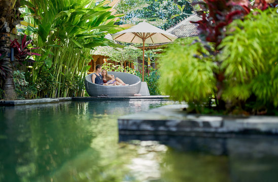 Candid Shot Of Young Caucasian Couple Lounging In Modern Chair Near Luxurious Pool Of Tropical Hotel And Spa In Bali