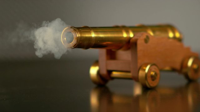 SLOW MOTION, MACRO, DOF: Vintage brass toy cannon shoots a small cannonball across the room. Small round shot comes speeding out the shiny barrel of a miniature cannon standing on a smooth surface.
