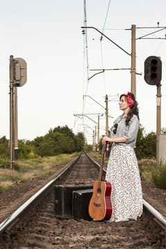 Beautiful girl with a guitar. Pretty woman with old suitcases on the train tracks. A teenager in a denim jacket, a long dress and a red bandage. Retro style