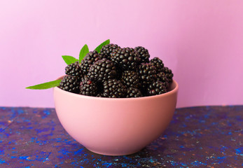  Berries of fresh blackberry in a plate decorated with mint leaves on a purple background.