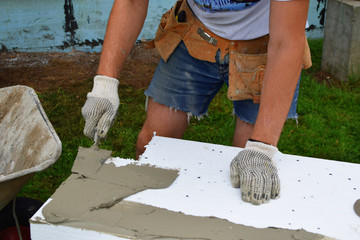Worker gloved hand with trowel applying glue on white rigid polyurethane foam sheet for house...