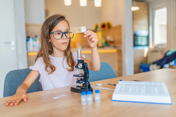 Confident little girl uses microscope at home. Beautiful elementary schoolgirl uses a microscope...