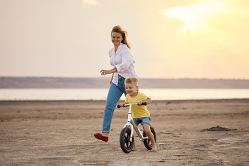 Mother with son riding bicycle in summer. Woman runs along with kid on the beach at sunset. Son...