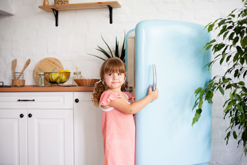 A little girl opens the door of the refrigerator. Beautiful kitchen.